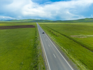 Fototapeta na wymiar Black passenger car is driving along an asphalt road along green fields, mountains on a sunny day. Photographing from a drone. Concept of travel