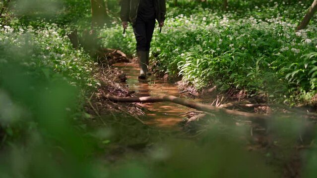 A man walking along a woodland stream surrounded by wild garlic while wearing wellies