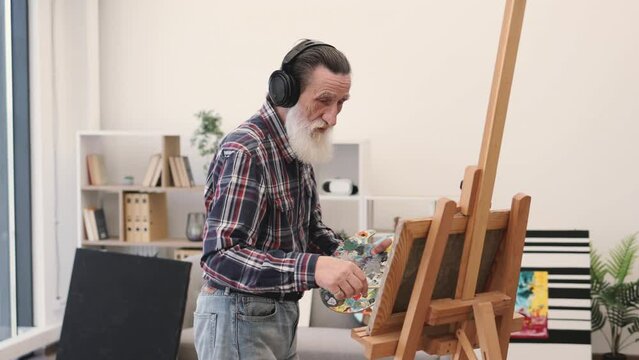 Cheerful elderly man in jeans using brush and palette while painting with audio accompaniment in headphones. Grey-haired professional expressing mastery through colors on canvas in art space.