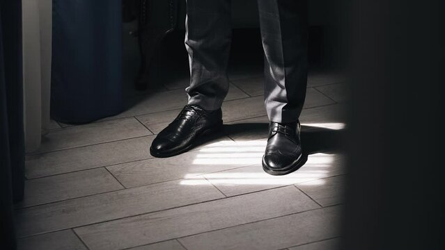 A man in trousers and black shoes stand in front of the window. His feet are illuminated by the sun's rays