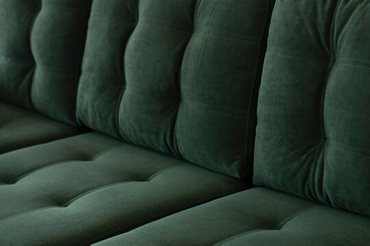 Soft vintage sofa in green velor with a quilted seat and a back of stitched cushions. Part of stylish furniture covered with plush fabric in emerald color close-up.