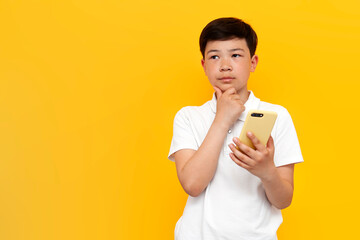 pensive asian little boy of ten years old in white t-shirt uses smartphone and thinks on yellow background