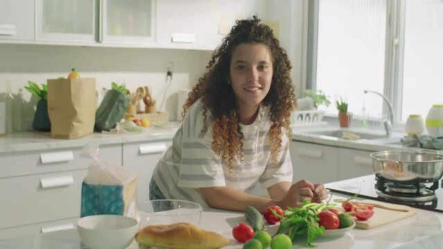 Medium shot of cheerful curly-haired girl leaning on kitchen table with food ingredients and posing for camera with smile during day at home