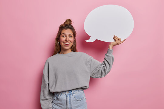 Horizontal shot of pretty young woman holds speech bubble ready to share her thoughts has eyes sparkle with anticipation inviting conversation sparking imagination dressed casually isolated on pink