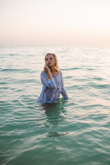 Attractive sensual woman standing in ocean water wearing wet shirt at beach over sunset outdoors. Summer vacation season. Looking at camera.