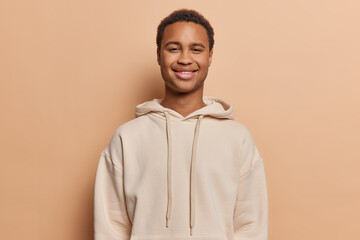 Studio shot of dark skinned pleased man smiles gladfully being in good mood during spare time dressed in casual hoodie isolated over brown background. Ethnic male teenager poses glad indoor.