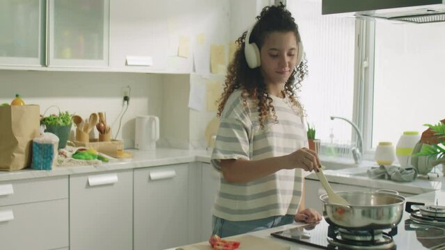 Medium long shot of cheerful girl in wireless headphones enjoying music while cooking food in kitchen at home