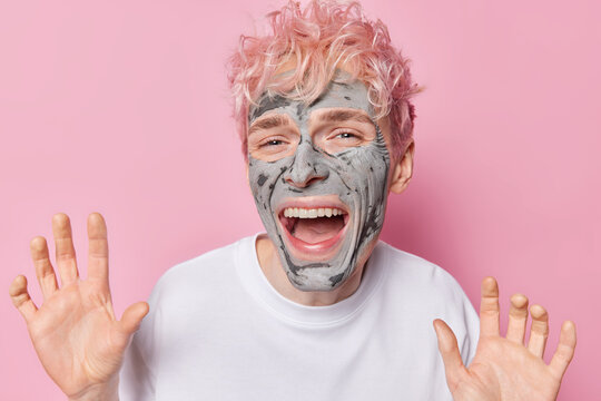 Photo of positive stylish pink haired guy exclaims loudly keeps palms raised up applies beauty clay mask on face for skin treatment dressed in casual white t shirt isolated over rosy background