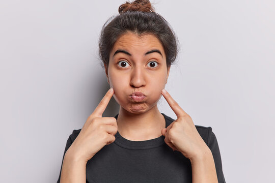 Portrait of funny young Brazilian woman with dark hair points at blowing cheeks stares surprisingly at camera wears casual black t shirt isolated over white background. Human facial expressions
