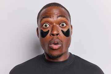 Photo of shocked dark skinned adult man applying hydrogel black beauty patches under eyes for wrinkle reduction engages in wellness procedures keeps lips rounded reacts to something terrible
