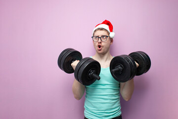 young nerd guy in glasses and santa hat trains and lifts heavy dumbbells on pink isolated background