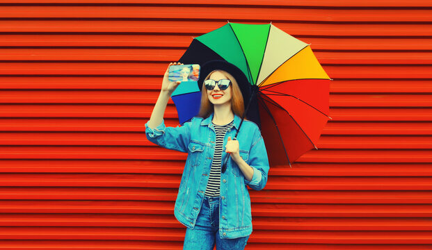 Portrait of happy smiling young woman taking selfie with phone with colorful umbrella on red background
