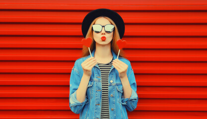 Portrait of beautiful young woman with lollipop blowing her lips with lipstick sending sweet air kiss wearing sunglasses, black round hat on red background