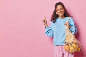 Eco living and zero waste. Positive curly haired woman carries fruits and vegetables in net bag points index finger on copy space tells about reasonable consumption isolated over pink background