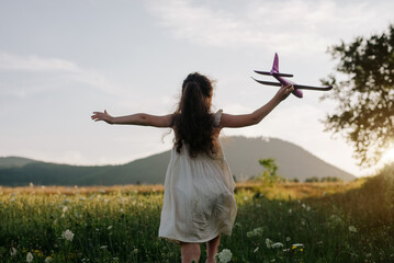 Back view of playful little girl kid run with airplane on background amazing mountains during summer warm sunset. Dream freedom concept. Child runs on field holding in hands toy aircraft