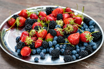 Ripe and appetizing blackberry, blueberry and strawberry in a serving plate on a wooden table. Natural light - 627305369