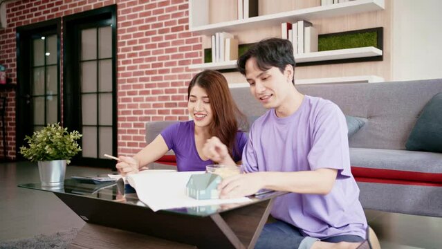 Young happy loving couple talking and using calculator to calculate the price of buying a house with a small wooden house model on a house drawing. Buying new home concept.
