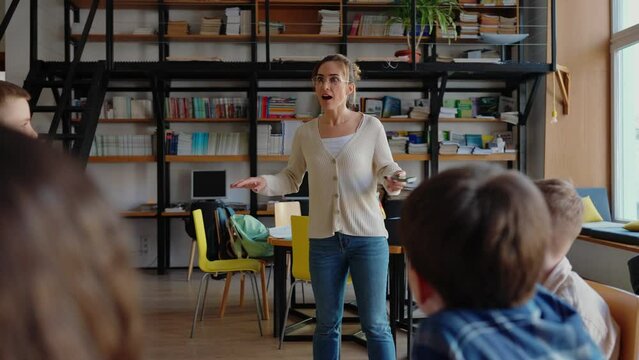 Handsome teacher explains rules of card game to children while sitting in circle on chairs in library at school