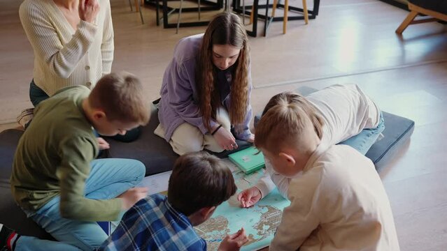 Serious children and their teacher look at map of world sitting on floor in library 
