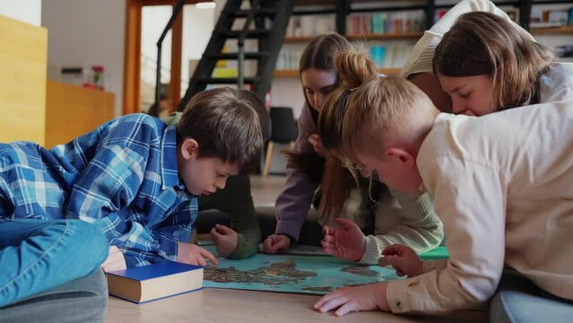 Concentrated children and their teacher mark points on world map with coin sitting on floor in library 