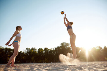 Two women, friends playing beach volleyball outdoors on warm summer day on sunset. Spending time in...