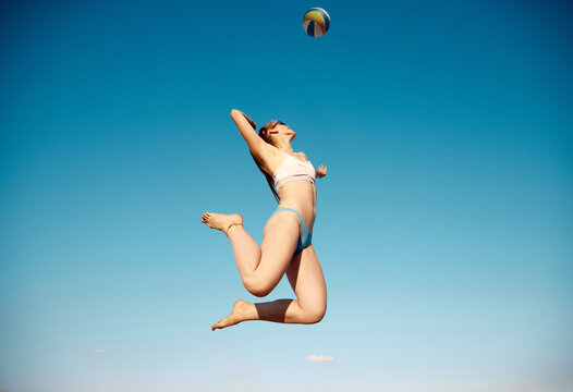 Bottom view dynamic image of young woman in swimsuit serving ball, playing beach volleyball over blue sky background. Concept of sport, active and healthy lifestyle, hobby, summertime, ad