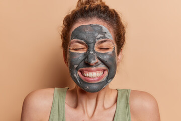 Studio shot of cheerful woman showcases her flawless smooth skin poses with clay beauty mask on...