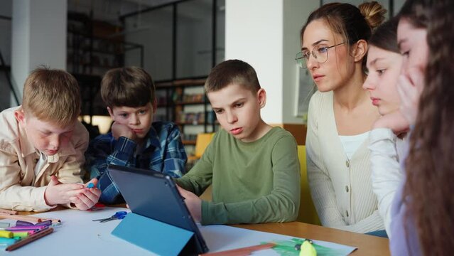 Serious teacher and children watching video on tablet at drawing table at library
