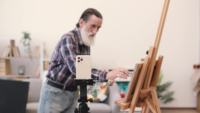 Elderly male painter with long grey beard using mobile phone with tripod for shooting video for family archive. Senior man dressed in jeans and checkered shirt demonstrating drawing skills on camera.