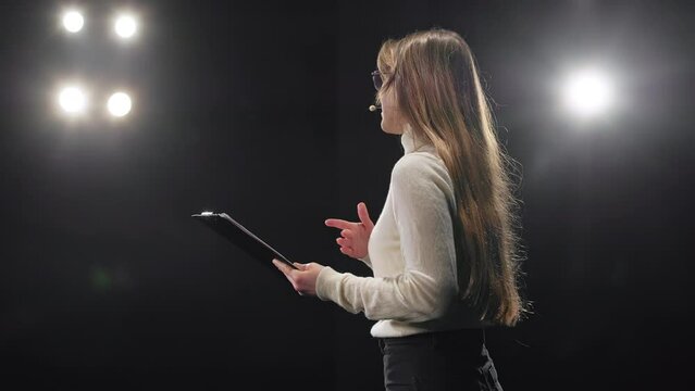Successful young businesswoman with clipboard in hands talking in microphone during motivational speech on stage. Caucasian blond advisor sharing her experience at modern workshop.