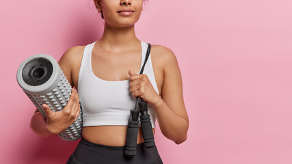 Cropped shot of unknown woman poses with sport equipment carries foam roller dressed in white top ready for yoga practice or training in gym poses against pink background with copy space for promo