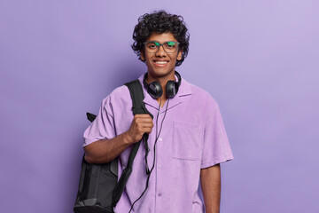 Cheerful millennial Hindu student poses over purple background dressed casually smiles pleasantly being on way to university with rucksack uses headphones for audio lessons. Studying and education
