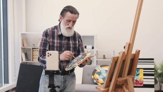 Aged man on retirement in casual outfit painting with brush on canvas using easel and looking straight at camera on smartphone set on tripod. Talented artist shooting video content about art.