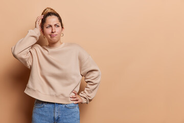 Studio shot of pensive young woman scratches head stands thoughtful considers something dressed in...