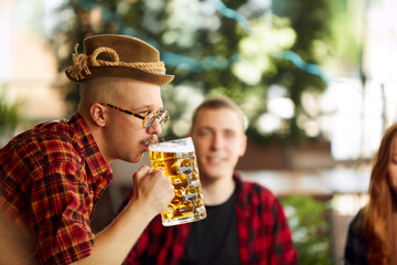 Close-up of young man in checkered shirt and fedora hat drinking beer. Blurred people in background. Sunny day. Concept of oktoberfest, traditional taste, friendship, leisure time, enjoyment