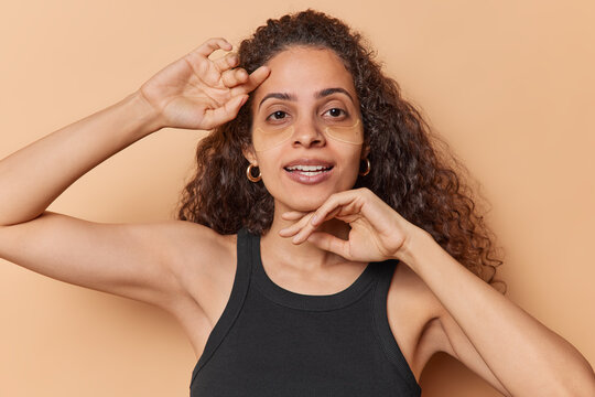 Photo of tender Brazilian woman touches face gently puts on hydrogel patches under eyes to reduce wrinkles wears casual black t shirt enjoys self care procedures isolated over brown background