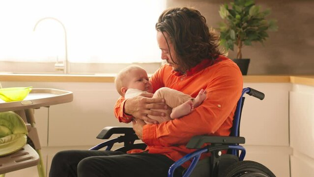 Portrait of young father in wheelchair lulling lovely toddler after breakfast in kitchen. Person with disabilities. Love, care. Family concept. Fatherhood. Paternity