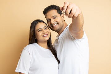 Happy young indian couple holding keys of newly purchase house flat or apartment isolated in beige background, Buying Real estate, Mortgage loan,Homeowners concept. Selective focus.