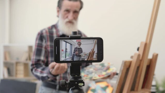 Blurred senior blogger broadcasting while painting new beautiful picture on easel at cozy art studio. Imposing elder male dressed in jeans and checkered shirt communicating with followers on internet.