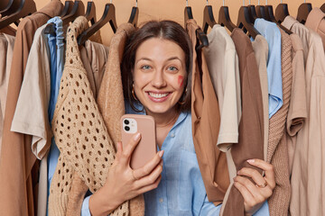 Glad young woman holds smartphone captivated by enticing display of incredible offer surrounded by clothes on hangers eagerly explores shopping mall determined to take advantage of big discounts