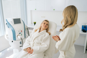 Back view of unrecognizable cosmetician consultation woman in white bathrobe lying on couch before rejuvenation cosmetology procedure in beauty clinic. Concept of anti-age rejuvenation skincare.