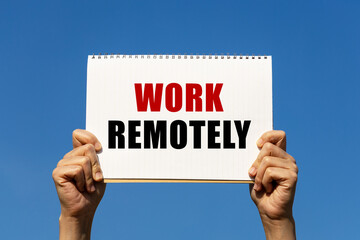Work remotely text on notebook paper held by 2 hands with isolated blue sky background. This message can be used as business concept about working remotely.
