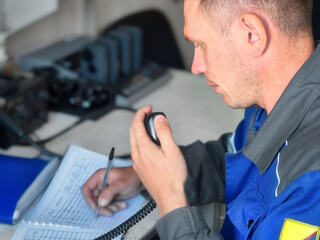 Man of Caucasian appearance receives signal on walkie-talkie and writes message in journal. Receiving distress call or important message via radio. Dispatcher sends coordinates.