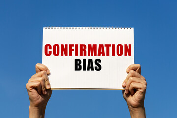 Confirmation bias text on notebook paper held by 2 hands with isolated blue sky background. This...