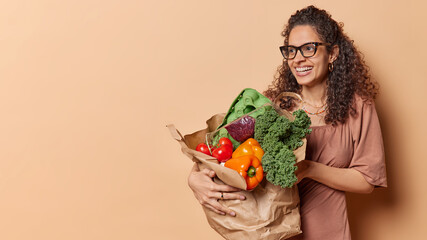 Young smiling Brazilian cheerful vegetarian woman in casual dress holding paper bag with vegetables...