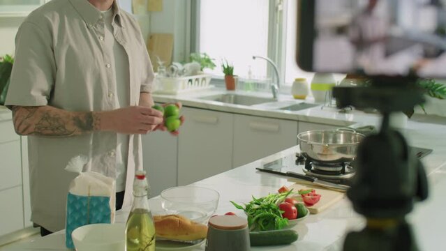 Medium shot of young man looking at smartphone camera and telling about food ingredients while giving online culinary masterclass or filming vlog in kitchen