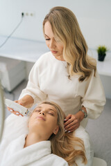Vertical portrait blonde woman client lying with closed eyes and getting stimulating beauty facial treatment during rf-lifting procedure. Concept of anti-age rejuvenation skincare.