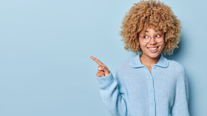 Horizontal shot of cheerful blonde curly young woman points index finger aside drawing attention to...