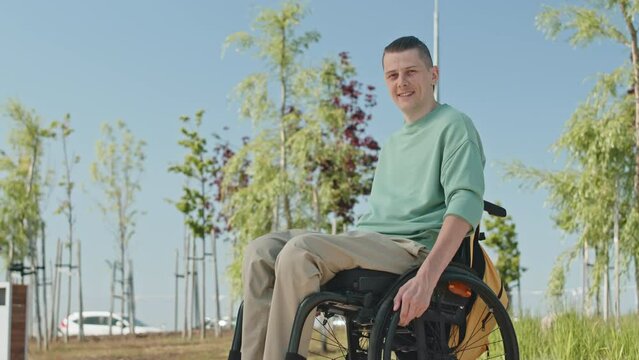Portrait of smiling young Caucasian man with disability posing for camera outdoors sitting in wheelchair under clear blue sky