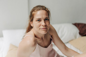 Sunny morning, a young woman with natural authentic beauty sitting on the bed looks at the camera.